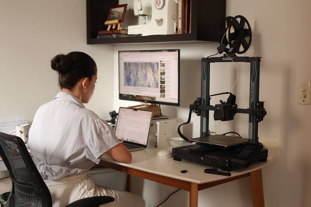 A person working at a laptop beside a 3D printer in a home office