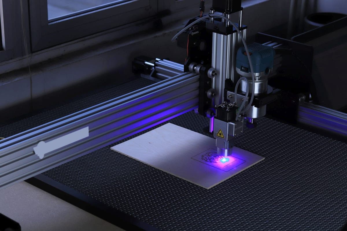 A 3D printer beside a desktop laser cutter in a workshop with dark walls