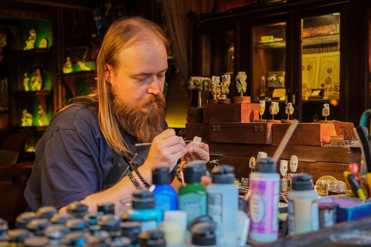 A hobbyist painting miniature models at a cluttered workbench surrounded by pots of paint